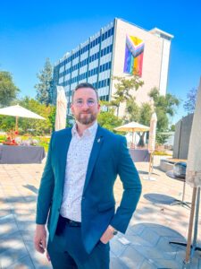 Dr. James Keane wears a blue shirt under a blue suit and glasses, posing in front of an office building at the NASA Jet Propulsion Laboratory with a multi-story Inclusive Pride Flag hanging from the size.