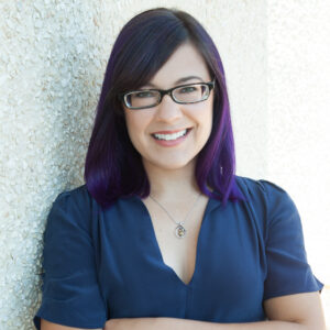 Liz Kruesi wears a blue blouse and glasses, has purple hair, and looks at the camera.