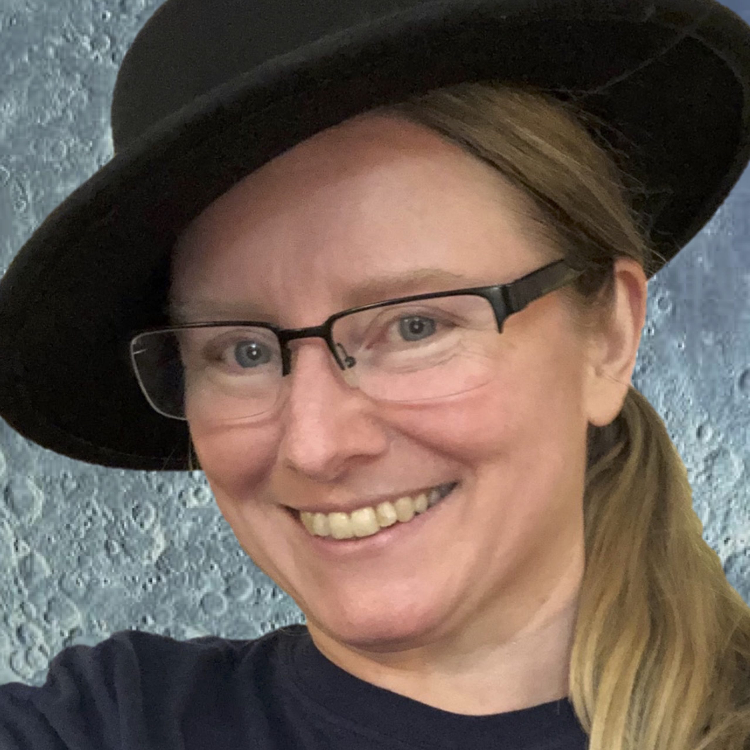 A white non-binary person wearing glasses and a black hat, with long blonde hair, smiles at the camera in front of a cratered lunar surface