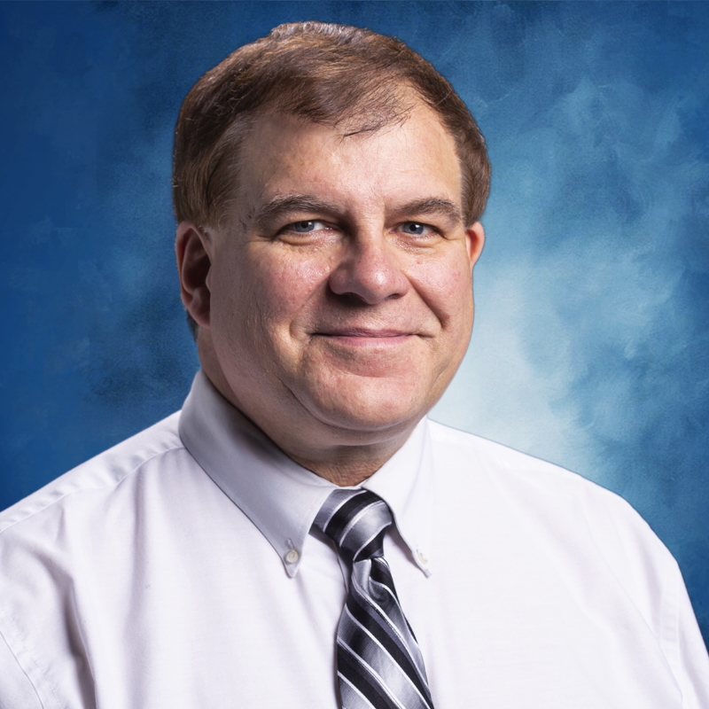 a white man wearing a light pink shirt and a blue & silver striped tie looks at the camera with a blue portrait background