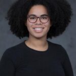 Hannah Wallace, a Black woman, wearing red-rimmed glasses with dark brown curly hair worn naturally and a black sweater, looks at the camera and smiles.