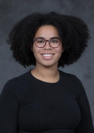 Hannah Wallace, a Black woman, wearing red-rimmed glasses with dark brown curly hair worn naturally and a black sweater, looks at the camera and smiles.